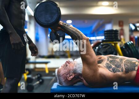 Senior fit man weight lifting with personal trainer in gym sport club - Mature bodybuilder doing workout session Stock Photo
