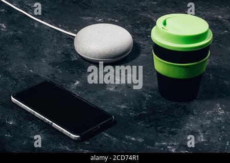 Digital Assistant concept. Smart speaker with LED lights activated on concrete black table with smartphone and reusable coffee cup. Stock Photo