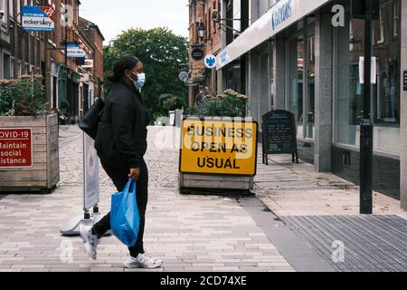 Black women in face mask with shopping bag walking along the main shopping street in Preson with 'Business Open as Usual' sign in background Stock Photo