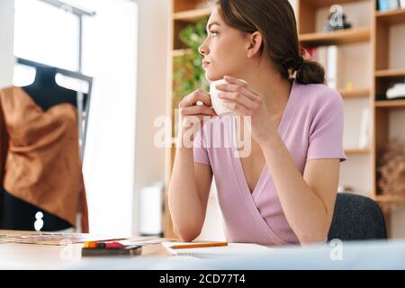 Photo of young nice woman fashion designer making sketches while drinking coffee at table in bright atelier Stock Photo