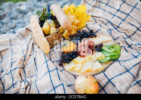 Outdoor picnic with a plate of meat and cheese cuts and grapes, a basket with a baguette and a bottle of wine on a mat. Stock Photo