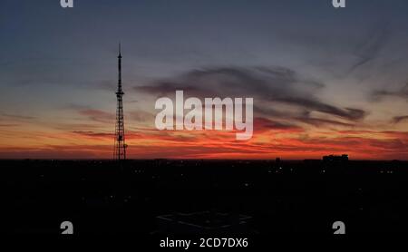 Blurry landscape of breathtaking sunset over urban skyline with communication tower silhouette. Purple and red clouds on dark evening sky background Stock Photo
