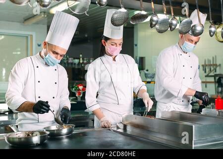 Chefs in protective masks and gloves prepare food in the kitchen of a restaurant or hotel. Stock Photo