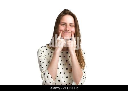 A young woman with long brown hair and a white polka-dot dress. she puts a smile on her face with both hands trying to make herself happy by smiling Stock Photo