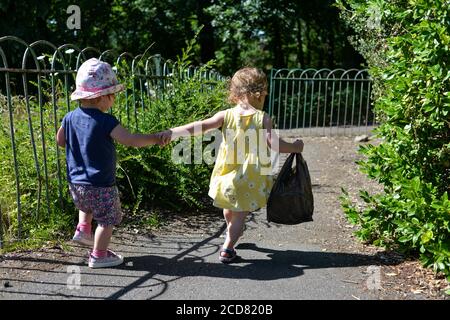 Two young children holding hands and walking away down a footpath in summer clothes and sunshine Stock Photo