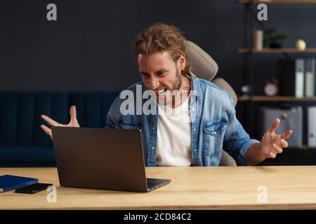 Angry young handsome curly man using laptop. Stressed freelancer lost data on broken not working laptop. Receiving bad news or email, spam message Stock Photo