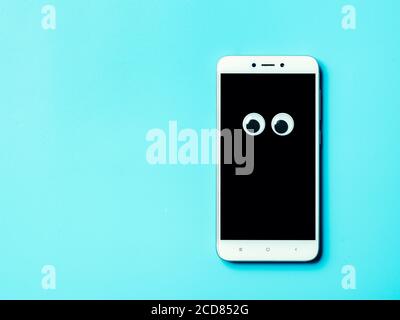 googly eyes on smartphone screen over blue background with copy space left. Top view or flat lay. Plastic toy eyes on dark phone screen background - halloween gadgets sale concept Stock Photo