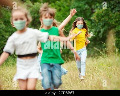 Happy little kids wearing protective face mask jumping and running on meadow, forest. Looks happy, cheerful, sincere. Copyspace. Childhood, pandemic concept. Healthcare, coronavirus pandemic. Stock Photo