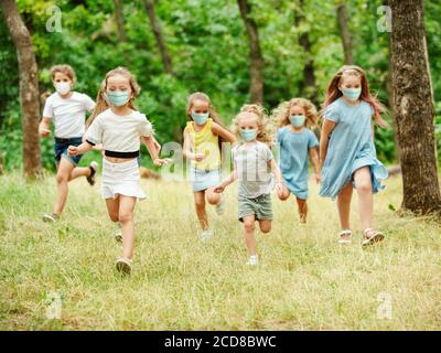 Happy little kids wearing protective face mask jumping and running on meadow, forest. Looks happy, cheerful, sincere. Copyspace. Childhood, pandemic concept. Healthcare, coronavirus pandemic. Stock Photo