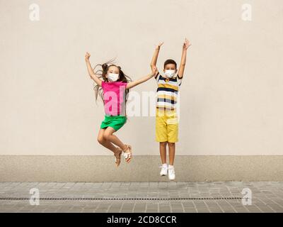 Happy little kids wearing protective face mask jumping and running on city street. Looks happy, cheerful, sincere. Copyspace. Childhood, pandemic concept. Healthcare, coronavirus pandemic. Stock Photo