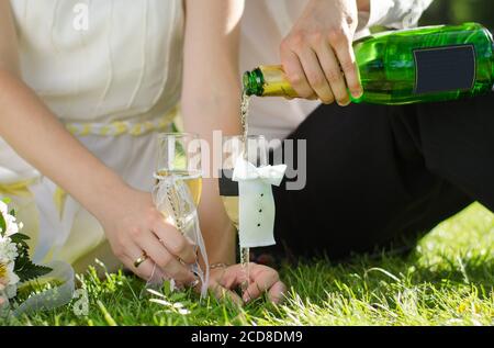 Wedding glasses stand on the grass, the groom's hand pours champagne into the glasses. Close up Stock Photo