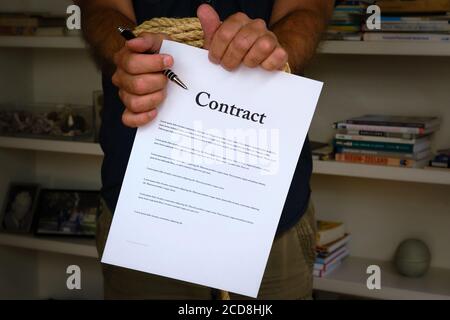 Tied hands holding pen and tearing contract paper in front of bookcase. Bound man hands rip paper contract, breach of contract. Unfair agreement Stock Photo