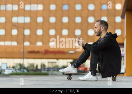 Young hipster man in casual black clothes using smart phone, resting, sitting on longboard or skateboard outdoors, side view Stock Photo
