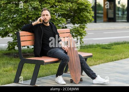 Young man resting, sitting with skateboard or longboard on wooden bench, talking on smart phone. Stock Photo