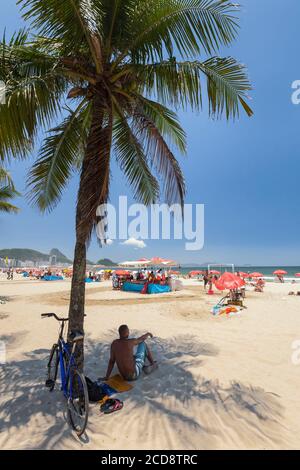 Brazil, Rio de Janeiro state, Rio de Janeiro city, Copacabana beach, Carioca landscapes between the mountain and the sea classified UNESCO World Heritage Site, man sitting on the sand, in a coconut tree shadow and looking the beach Stock Photo