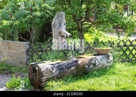 Wooden water bowl and water tap emerging from a chopped tree trunk, carved in the shape of a face. Stock Photo
