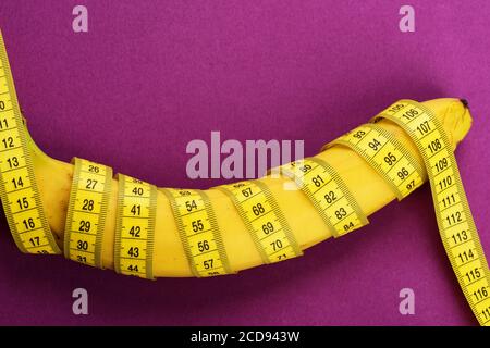 Yellow tape around banana isolated on violet background. Slim body and healthy food concept. Banana with yellow tape for measuring figure. Rolled centimeter ruler spinned around fresh fruit. Stock Photo