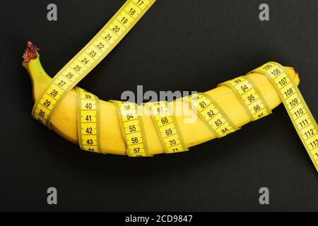 Weight loss, diet and healthy lifestyle concept. Centimeter ruler spinned around fresh fruit. Banana with yellow tape for measuring figure. Yellow tape around banana isolated on black background. Stock Photo