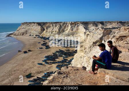 Morocco, Western Sahara, Dakhla, young men sitting above the cliff overlooking Araiche beach and fishing boats Stock Photo