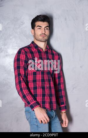 A serious handsome young man in a red checkered shirt standing in front of a grey wall in a studio. Stock Photo