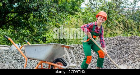 girl with wheelbarrow of rubble. kid working on construction site. teen girl takes out rubble from wheelbarrow. kid with shovel loading crushed stones. Laying the foundation, building project. Stock Photo