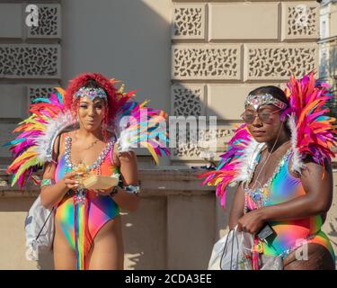 Two women in colorful costumes, one of which is eating, seen standing near a building before the start of the Notting Hill Carnival parade 2019. Stock Photo