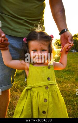 Cute little girl take hands with her father. Walking with kids concept image. Little girl holding hands with her parents. Smiling baby in dress. Stock Photo