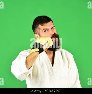 Japanese martial arts concept. Combat master hits himself with fist. Man with beard in white kimono on green background. Karate man with suffering face in uniform and golden boxing gloves. Stock Photo