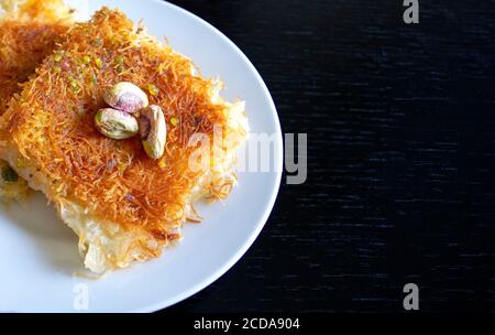 Traditional eastern dessert kunafa. Top view. Arabic cheese kunafa in white plate on dark wooden table with copy space. Konafa. Stock Photo