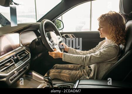 attractive curly caucasian child girl sit inside of new car, she likes this car, want parents to make purchase in dealership