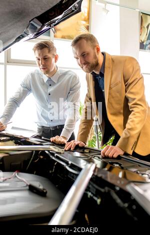 young caucasian cars salesman show the perfect assemble hood of car in dealership. business man examining all details, want to buy Stock Photo