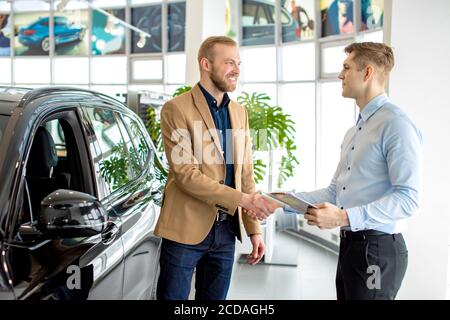 happy caucasian man owner of new car, stand in dealership shake hand to salesman. successful purchase in dealership Stock Photo