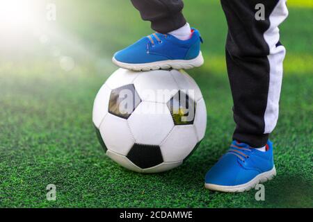 Children's foot of the winner in sports shoes sneaker stands on a soccer ball against a background of grass. Close-up street shot of game and training Stock Photo