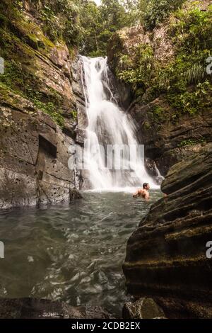 A tourist swims in the pool below La Mina Falls or Cascada la Mina in the rainforest of El Yunque National Forest in Puerto Rico. Stock Photo