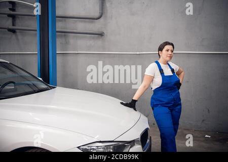 Car service worker in blue overalls leans on the hood of the car and poses Stock Photo
