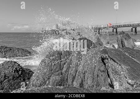 The much photographed Palm Cove jetty with a splash of red on a black and white image of a wave breaking on the rocks at the northern end of the beach. Stock Photo
