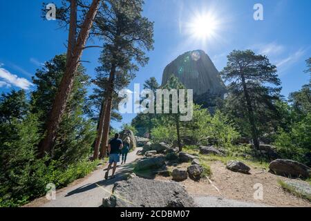 Wyoming, USA - June 23, 2020: Tourist hikers walk towards Devils Tower National Monument in the morning. Sunflare in photo Stock Photo