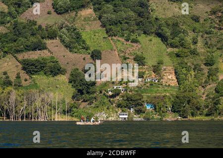 Mayan men paddle a cayuco or canoe on Lake Atitlan in front of houses and farm plots on the steep slopes of the San Pedro Volcano, near Santiago Atitl Stock Photo