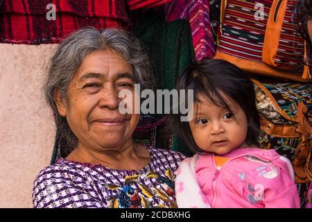 An older Tzutujil Mayan woman in traditional dress holds her little granddaughter in a shop in Santiago Atitlan, Guatemala. Stock Photo