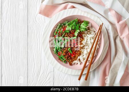 Asian minced pork and long grain rice with green beans stir fry sprinkled with sesame seed, chili pepper on top, served on a pink bowl on a white wood Stock Photo