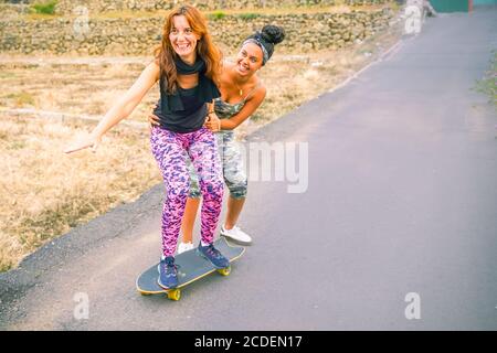 Young mother practising on skateboard in the street. Daughter helping mum on skateboard. Mum learning to ride skateboard as Daughter teaches her in th Stock Photo