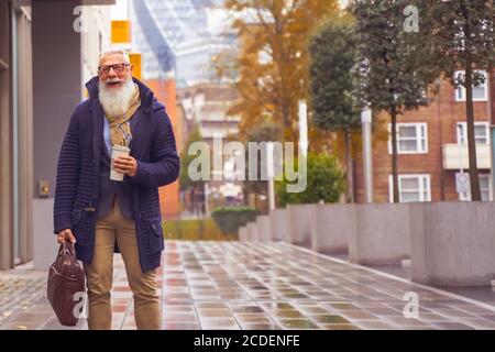 Happy senior businessman drinking coffee and leaving work. Man walking on a rainy day in the middle of autumn. work and fashion lifestyle concept - Fo Stock Photo