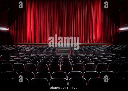 Darkened empty movie theatre and stage with the red curtains drawn viewed over rows of vacant seats from the rear Stock Photo
