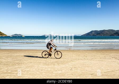A man wearing a face mask as a preventive measurerides a bicycle at Acores Beach. Stock Photo