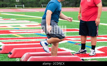 A high school football lineman is running an agility drill over red barriers ahile the coach is watching on a turf field. Stock Photo