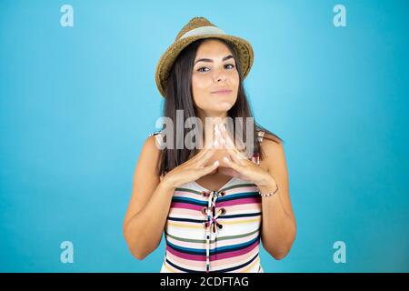 Young beautiful woman wearing swimsuit and hat over isolated blue background Hands together and fingers crossed smiling relaxed and cheerful. Success Stock Photo