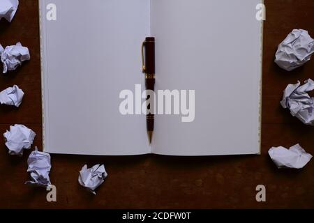 Closeup and selective focus on blank notebook with pen placed on wooden table surrounded by crumpled paper Stock Photo