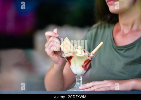 Close up of a delicious looking ice cream cup with an out of focus woman grabbing a spoonful. Shot taken from outside. Ice cream and lifestyle concept Stock Photo