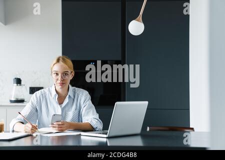 Photo of focused young woman in eyeglasses working with cellphone and laptop while sitting at table in kitchen Stock Photo