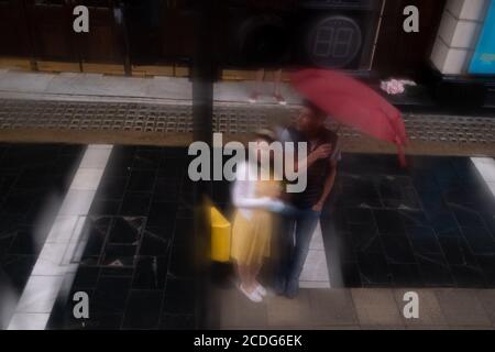 Seen through a wet bus window, a man holds a red umbrella over himself alongside a woman during summer rains on Shaftesbury Avenue, on 27th August 2020, in London, England. Stock Photo
